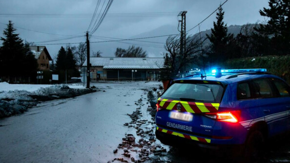 Un véhicule gendarmerie, vu de trois-quarts arrière, gyrophare allumé, stationné en bordure d'un chemin inondé, à la nuit tombante. En arrière-plan, un fleuve d'eau boueuse dévale au milieu des habitations.
