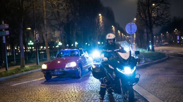 Un gendarme en moto de nuit devant des véhicules de collection