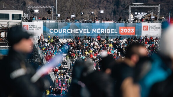 En amorce sur l'image se trouve en flou des personnes, dans le fond, sur les gradins, se trouve le public avec noté dans le fond, "Courchevel -Méribel 2023"