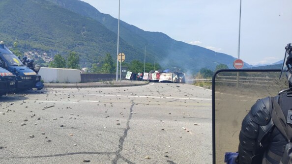 Au premier plan, à droite, un gendarme mobile vu de dos, portant un bouclier. A gauche, l'avant de deux Irisbus et plusieurs gendarmes mobiles. Face à eux, sur la route, laquelle est jonchée de pierres et autres projectiles, un groupe de manifestants radicaux.