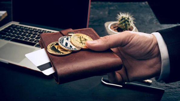 Photo d'illustration représentant un homme tendant un portefeuille avec des grandes pièces de monnaie, devant un bureau sur lequel est posé un ordinateur.