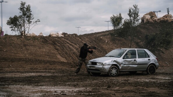 Un plastron armé d'un FAMAS lors d'une démonstration, debout et protgé par un véhciule épave gris, s'apprête à tirer vers la droite. Derrière lui, un talus