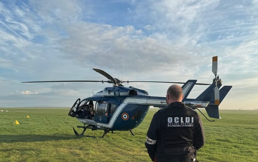 Un homme vêtu d'un uniforme noir portant dans le dos la mention blanche OCLDI se tient debout, de dos, au premier plan. Il regarde un lélicoptère bleu de la gednarmerie nationale, dont la porte est ouverte, posé sur une vaste pelouse verte.  Le ciel bleu est parsemé de nuages gris et blancs.