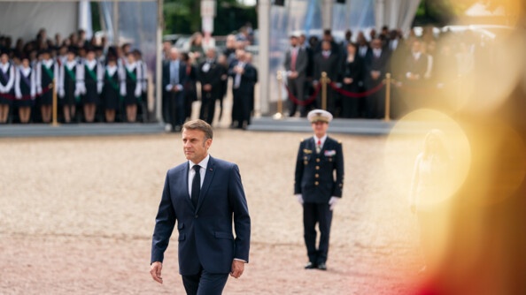 Le président de la République française avance dans la cour du Mont-Valérien. Derrière lui, se dessine en flou toutes les autorités présentes. Au milieu de la cour également derrière lui se trouve un militaire.