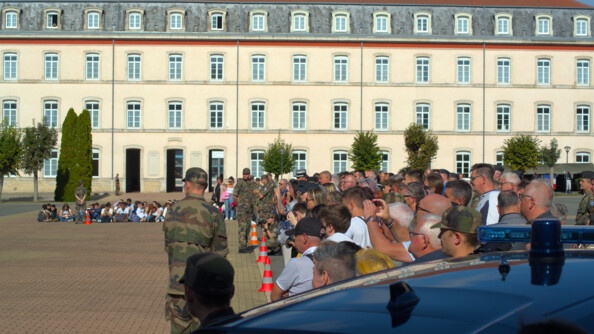 Devant un grand bâtiment de couleur clair se trouvent des jeunes assis par terre. A droite se trouve du public qui regarde dans la même direction. En tout premier plan se trouve le toit d'une voiture de gendarmerie avec un girophare étient. De dos se tient debout un militaire en tenue kakie