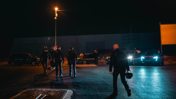Cinq gendarmes marchent de dos dans la rue. Ils sont éclairés par les phares d'une voiture de gendarmerie.