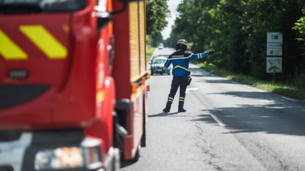 De dos sur la route un motocycliste de la gendarmerie est en train de faire la circulation. A gauche en flou un camion de pompier,