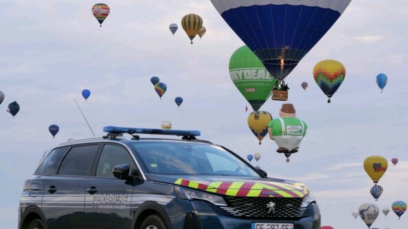 une voiture de la gendarmerie est au premier plan. Au second plan, les montgolfières prennent leur envol