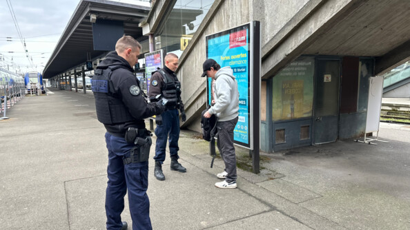 Deux gendarmes en train de contrôler un jeune homme vêtu d'un pantalon gris foncé, sweat gris clar et casquette noire dans une gare, sur les quais