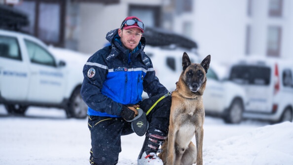 Sur une route enneignée, devant des véhicules blancs floutés, se tient accroupi un homme d'âge moyen, vêtu d'une tenue de ski bleue et d'une casquette rouge. Contre sa jambe gauche, est appuyé un berger blege malinois, assis et tenue en laisse par l'homme.