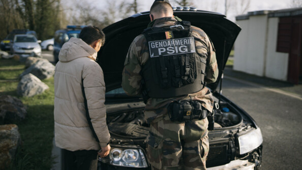 Un gendarme de dos ainsi qu'une personne interpellée. Ils sont entrain de contrôler l'intérieur du capot d'une voiture.