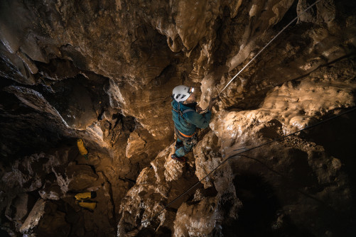 Photo montrant un gendarme spéléologue en combinaison et casque équipé d’une lampe frontale, en train d’escalader une paroi rocheuse à l’intérieur d’une grotte.