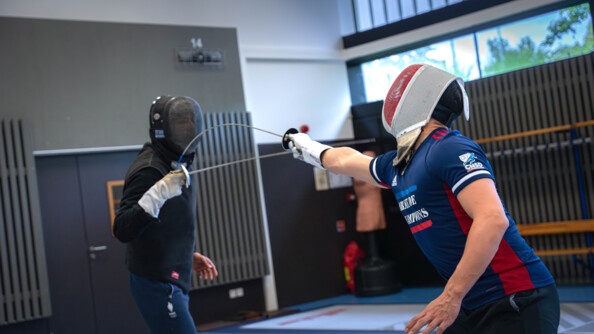 Un escrimeur vétu du survêtement de l'armée de champions touche son maître d'arme face à lui, vêtu de noir, qui ne pare pas