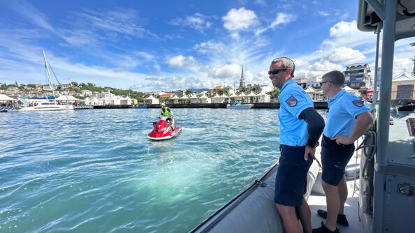 Deux gendarmes à droite sur un bateau et un gendarme en jet-ski en mer, devant un port.