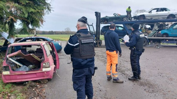 Un gendarme, un policier municipal, et un homme en pantalon de chantier orange, de dos, avec à leur gauche deux épaves de véhicule et au fond à droite un homme arrimant des voitures épaves sur un camion