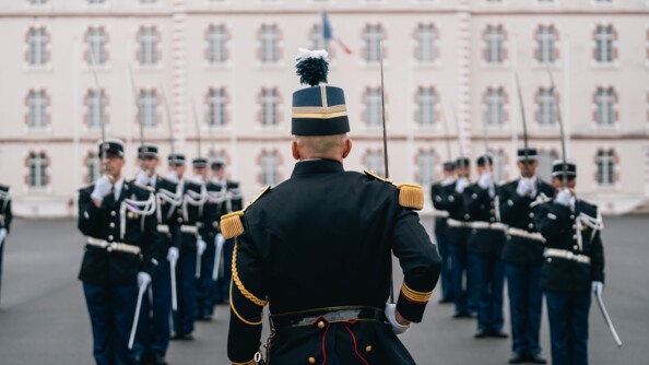 Des gendarmes en tenue de cérémonie sur 2 colonnes sur la place d'armes devant un bâtiment, au présentez sabre. Au milieu, de dos, un officier en tenue de tradition