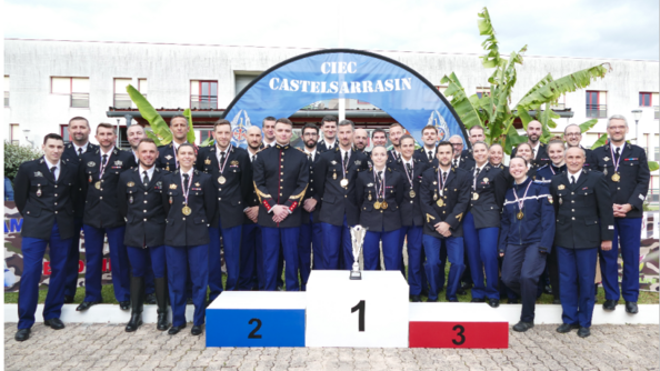 L'équipe de gendarmerie du championnat de France militaire de badminton 2024, composée de 32 personnes, en uniforme, pose, souriante devant le podium revêtant les couleurs du drapeau français. Sur la premère marche est posée la coupe rempprtée par l'équipe. Derrière eux, une grande bannière bleue en arc porte la mention "CIEC Castelsarrasin"