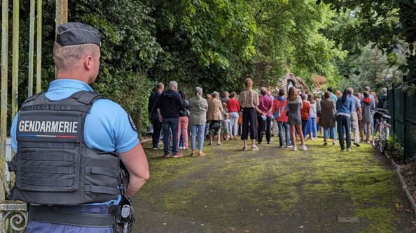 En extérieur, un gendarme en premier plan se tient debout et surveillent une cérémonie religieuse qui se déroule en second plan.