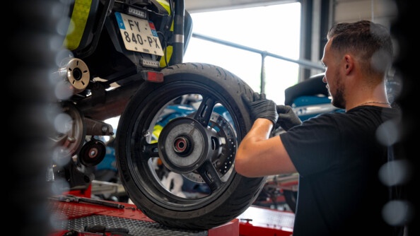 Un mécanicien, vêtu d'un t-shirt noir et de gants noirs, répare une moto bleue de la gendarmerie. Il se tient debout, de biais, et travaille sur le pneu arrière.