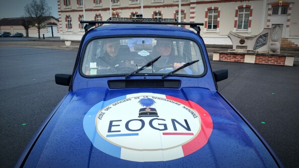 Deux hommes roulent &agrave; bord d'une Renaud 4L bleu marine, dont le capot est couvert d'un grand &eacute;cusson comportant la mention "Ecole des officiers de la gendarmerie nationale - EOGN". Ils circulent dans l'enceinte d'un b&acirc;timent, que l'on aper&ccedil;oit en arri&egrave;re plan. Des gouttes de pluie sont visibles sur le capot de la voiture.
