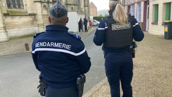 Deux gendarmes de dos au premier plan, à gauche un homme et à droite une femme, dans une rue. A gauche de la rue le mur d'un église, à droite la façade d'un bâtiment normand