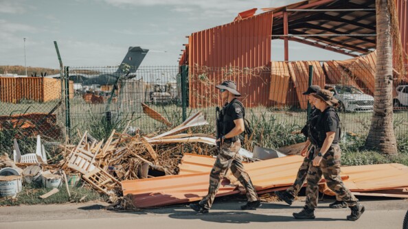 Trois gendarmes en traillis et haut noir le long d'une grille avec des tôles à demi-envolées; au fond à gauche le dos et la queue d'un avion