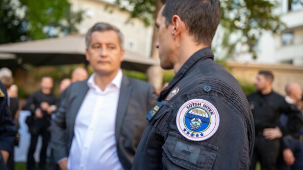Gérald Darmanin se tient debout, les mains sur les hanches, face à un homme que l'on aperçoit de biais, portant un uniforme avec  l'insigne du GIGN. Derrière eux, des personnes - dont l'image est floutée - discutent dans un jardin.