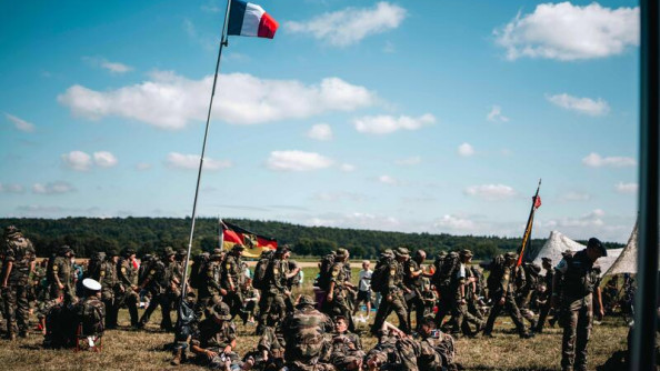 Un groupe de militaires, en treillis kaki, photographiés de biais, avacent sur une pelouse. Plusieurs drapeaux de différents pays émergent du groupe, dont un drapeau français.