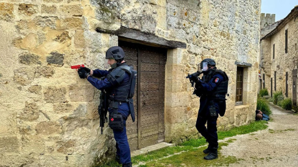 Deux gendarmes équipés en gilet lourd et avec des armes fictives rouge arrivent à un coin de mur