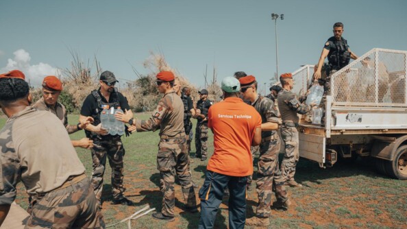 Des militaires et gendarmes ainsi qu'un personnel du service technique en t-shirt orange et pantalon bleu déchargent des bidons d'eau d'un pick-up blanc