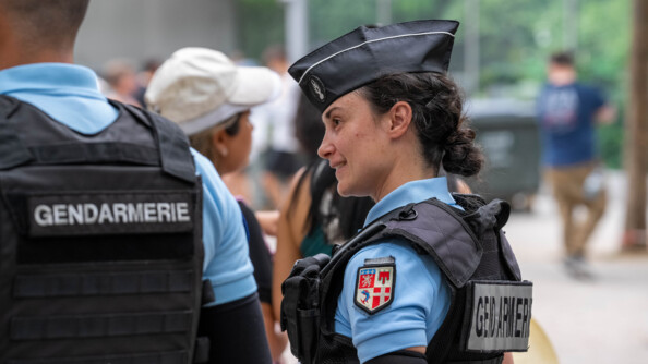 Une femme gendarme se tient ebout, de profil. Elle sourit. Elle est brune, porte un gilet pare-balles et une queue de cheval. A ses côté, se tient un second gendarme dont on aperçoit le dos. Autour, on distingue des passants, dont l'image est floutée.