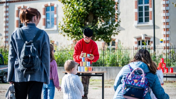 Deux femmes et trois enfants de dos devant des jeux de chamboule-tout sur des petites tabes. Une femme en pantalon de treillis, sweat rouge et bérêt noir, installe le jeu