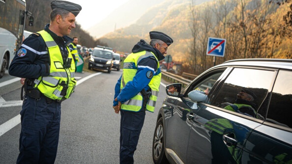 Deux gendarmes avec chasuble jaune conseillent un automobiliste sur la route. On peut voir sur la gauche une longue colonne de voitures.