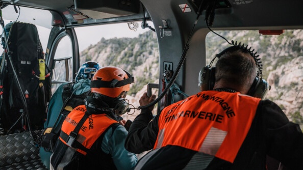 Trois hommes de dos dont deux avec des chasubles orange floquée en noir "secours en montagne gendarmerie" dans la caine d'un hélicoptère. Au fond en flou à travers els ouvertures de l'hélicopètre, la végétation des flancs de montagne