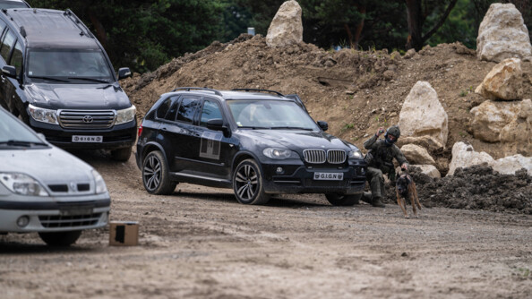 Devant deux 4x4 noirs, un ma&icirc;tre de chien du GIGN envoie son animal qui court enlui indiquant un point au laser
