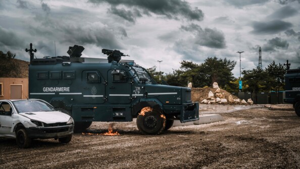 Un centaure de la gendarmerie avec un sol et la roue avant droite légèrement enflammée. A gauche un véhicule épave blanc