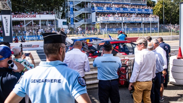Deux gendarmes de dos, entourés de nombreuses personnes. Tous regardent vers la piste devant eux où sont arrêtées deux voitures de course. De l'autre côté de la seconde barrière, les spectateurs