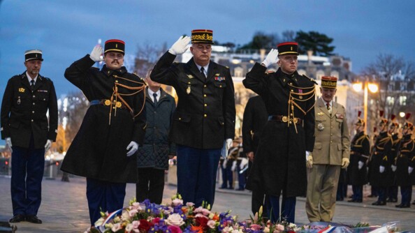 Au premier plan, un g&eacute;n&eacute;ral de gendarmerie, encadr&eacute; par deux gendarmes, tous trois en grande tenue et au garde-&agrave;-vous devant une gerbe de fleurs.