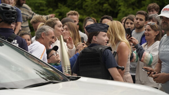 Derrière un véhicule, une gendarme mobile de profil, à sa gauche une homme portant la flamme olympique, le tout tèrs proche de la foule, tous prenant des photos et souriant