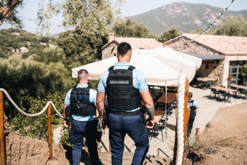 Deux gendarmes, dos à l'objectif, se dirigent vers une maison en pierres située en contrebas.