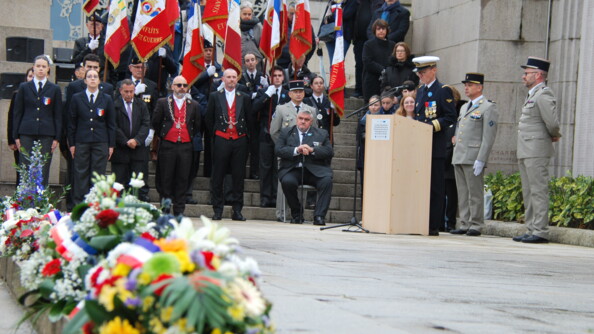 Sur un sol pavé on aperçoit une grande gerbe de fleurs colorées, tout en longeur. En arrière plan se tiennent des militaires. Plusiers d'entre eux portent des drapeaux. L'un d'eux, se tenant derrière un pupitre, équipé d'un micro, semble prononcer un discours