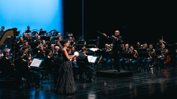 Une femme tenant un globe lumineux dans les mains avec une robe longue grise sur une scène au milieu de l'orchestre de la garde républicaine