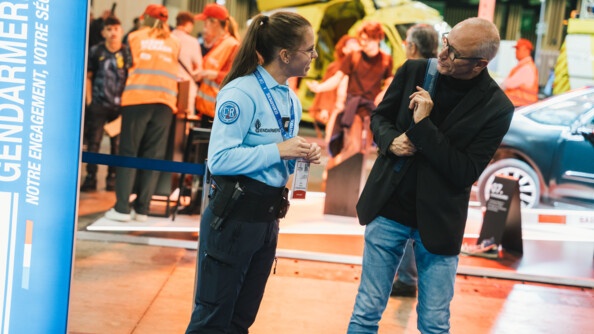 Une jeune femme gendarme, vêtue d'un pantalon bleu marine et d'un polo bleu ciel à manches longues, discute avec un visiteur sur le stand gendarmerie du Mondial de l'Auto de Paris 2024. Tous deux sont souriants.