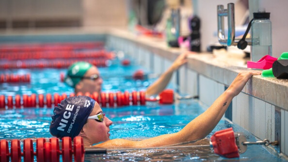 Une nageuse revêtant un bonnet bleu marine ainsi que des lunettes de nage, récupère, au bord du bassin, les mains posées sur le rebord de la piscine. Derrière elle, on aperçoit une autre nageuse, dont le visage est floutée,  qui porte un bonnet vert, et se tient dans la même position qu'elle.