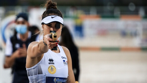 Une femme de face vétue d'un haut de sport blanc sans manche vise droit devant, son visage est caché par le pistolet, un oeil masqué par un cache et elle porte une casquette blanche