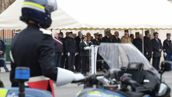 Au premier plan, flou, un motocycliste en grande tenue, vu de profil. En arrière-plan, focus sur la préfète qui lit l'ordre du jour. Derrière elle, un parterre d'autorités civiles et militaires.