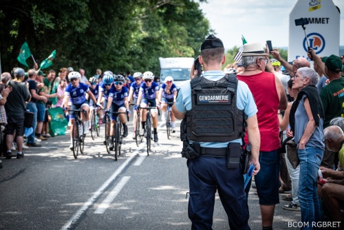 Un gendarme, vu de dos, surveille le passage d’un groupe de cyclistes féminines en tenue bleue pendant une course. Il porte un gilet pare-balles marqué « Gendarmerie ». Les spectateurs, massés des deux côtés de la route, applaudissent et prennent des photos. L’ambiance est animée et festive, dans un cadre verdoyant.