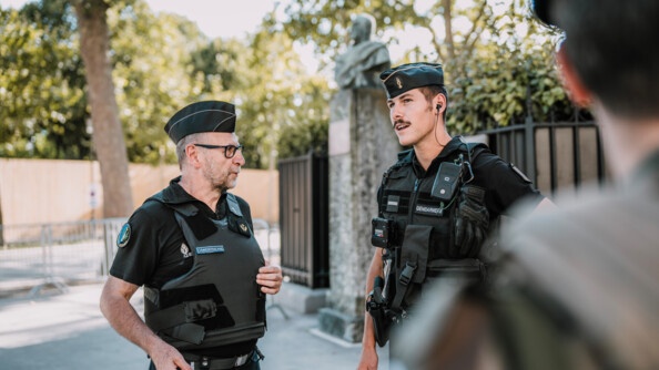Un aumponier de gendarmerie catholique en tenue noire avec un gilet pare-balle et porteur de lunettes, dans la rue, échanegant avec un gendarme mobile vêtu de noir en train de parler