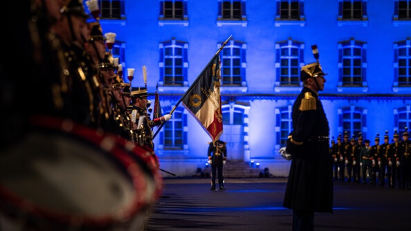 EN flou au premier plan les tambours de la Garde républicaine. AU second plan, net, le drapeau de l'EOGN. A droite, un garde républicain. En fond, l'Etat-major de l'EOGn illuminé en bleu et à droite, les élèves officiers alignés en grande tenue