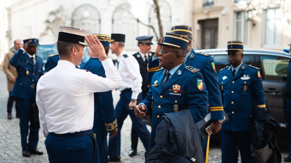 A l'extérieur, sur un sol pavé, se saluent des militaires de la gendarmerie congolaise et des militaires de la gendarmerie nationale, soit huit personnes au total. Tous portent le képi. En second plan, on aperçoit une voiture.